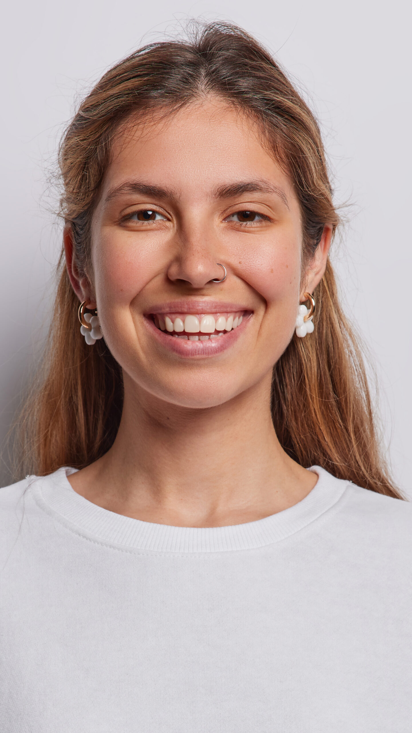 Portrait of cheerful long haired woman smiles toothily being in good mood dressed in casual sweatshirt has piercing in nose isolated over white background. People postive emotions and feelings concept Woman in white shirt with earrings looking at camera